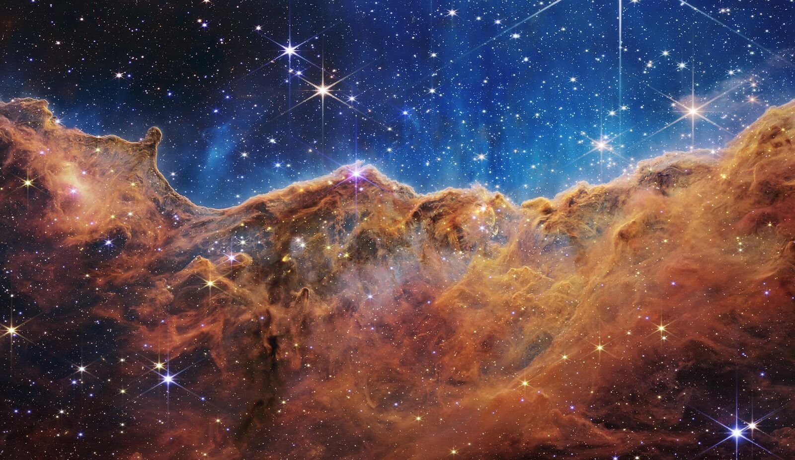 Penhascos de gás e poeira alaranjados com muitas estrelas ao fundo, na Nebulosa da Quilha